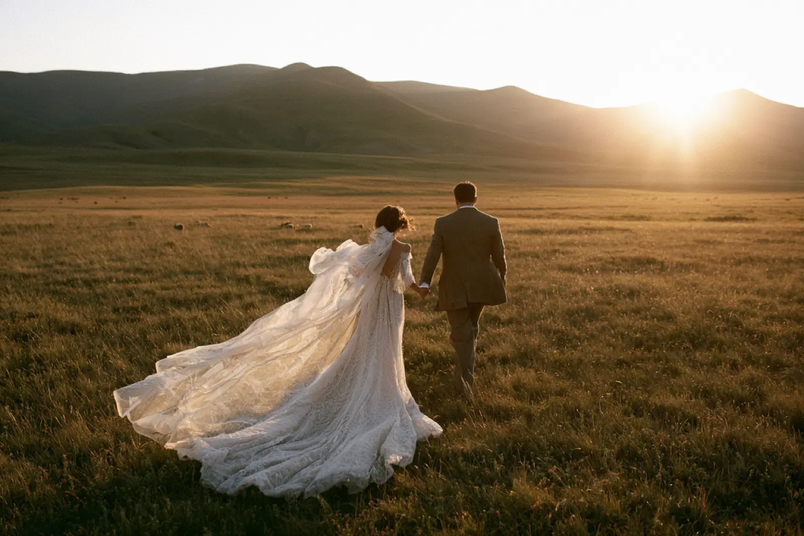 A bride and groom walking through a wide field toward the setting sun