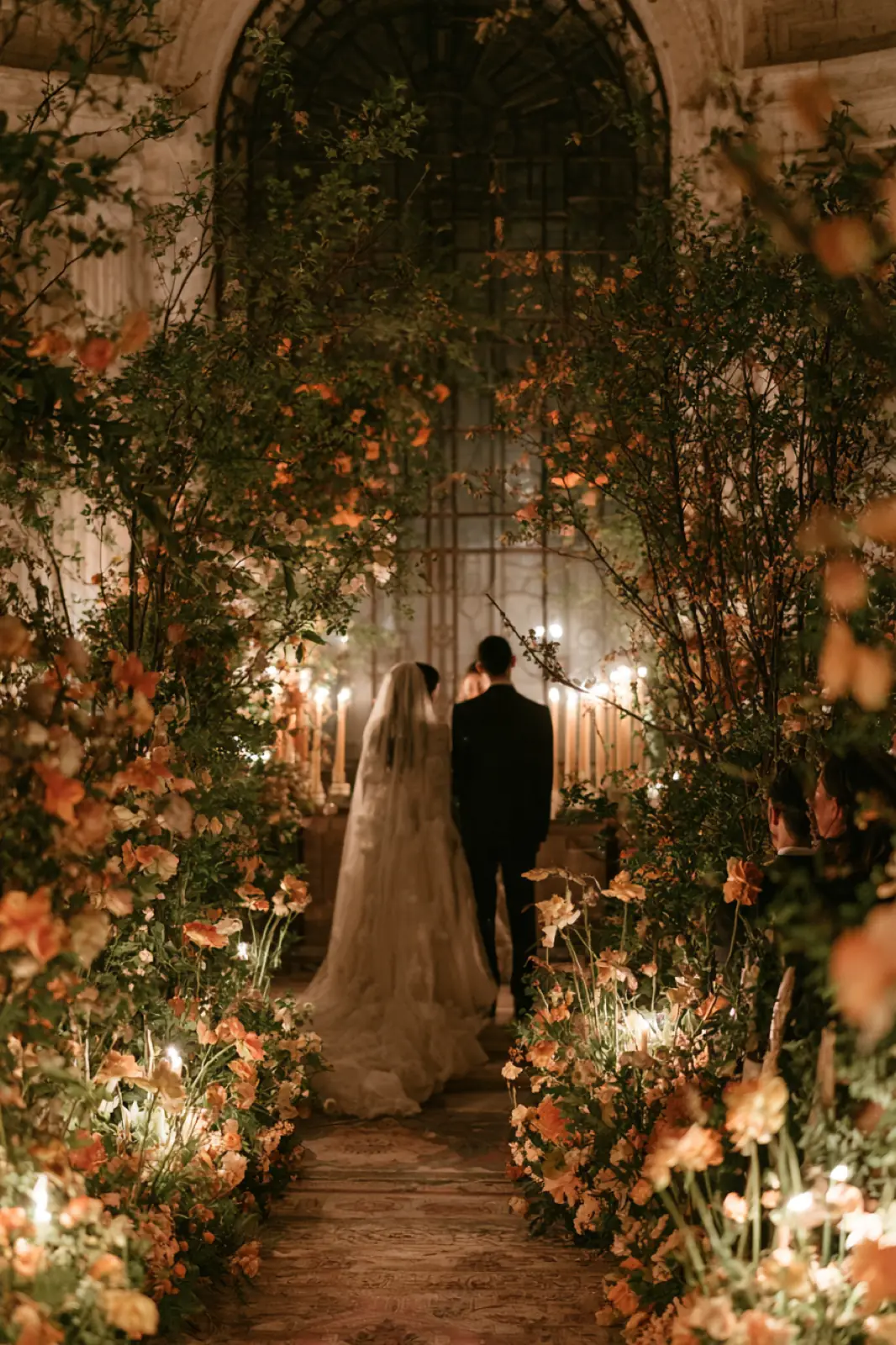 A couple heading off through an arch of flowers at dusk, her veil trailing behind