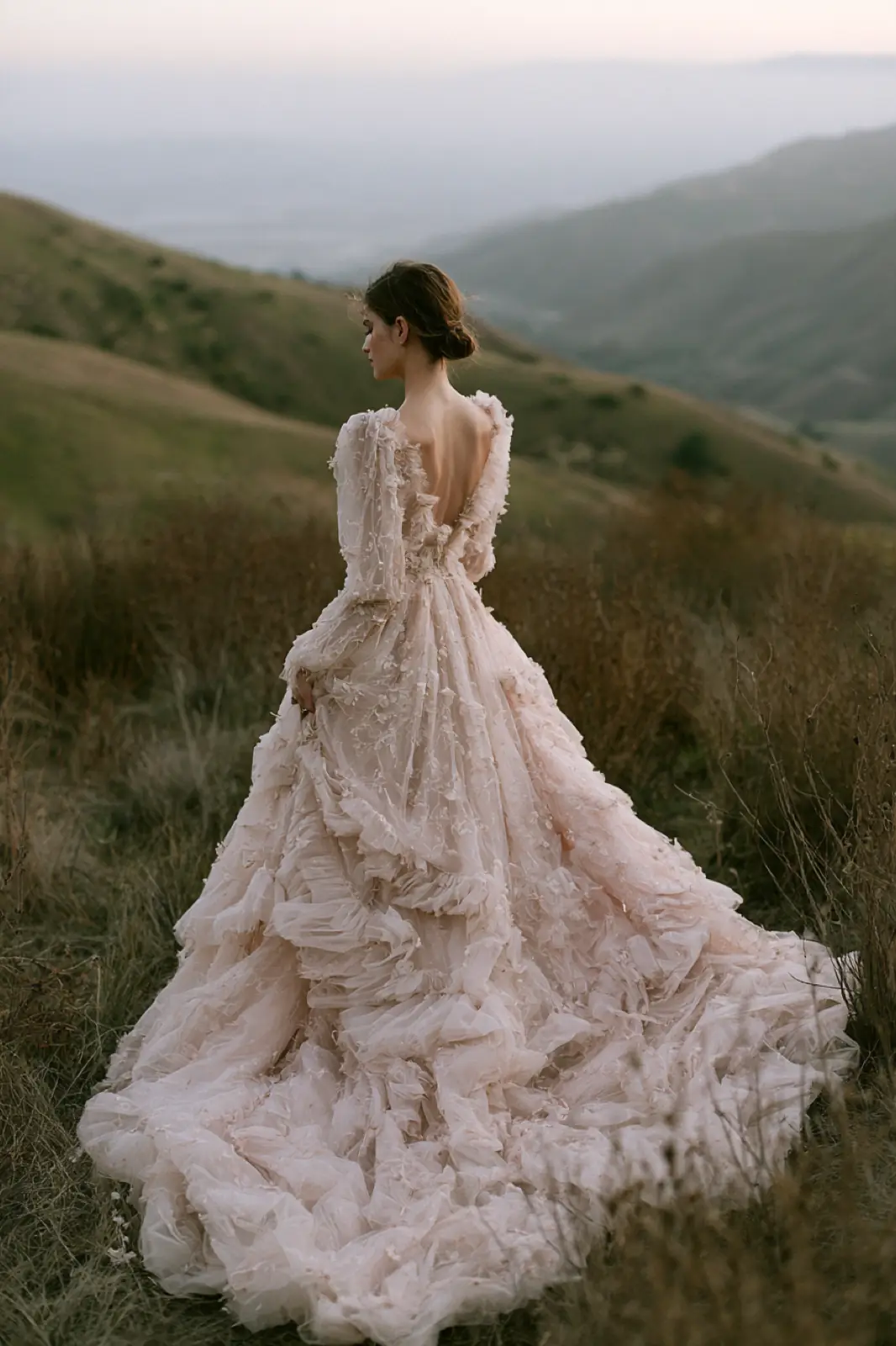 A bride in a full blush gown standing on a green hillside