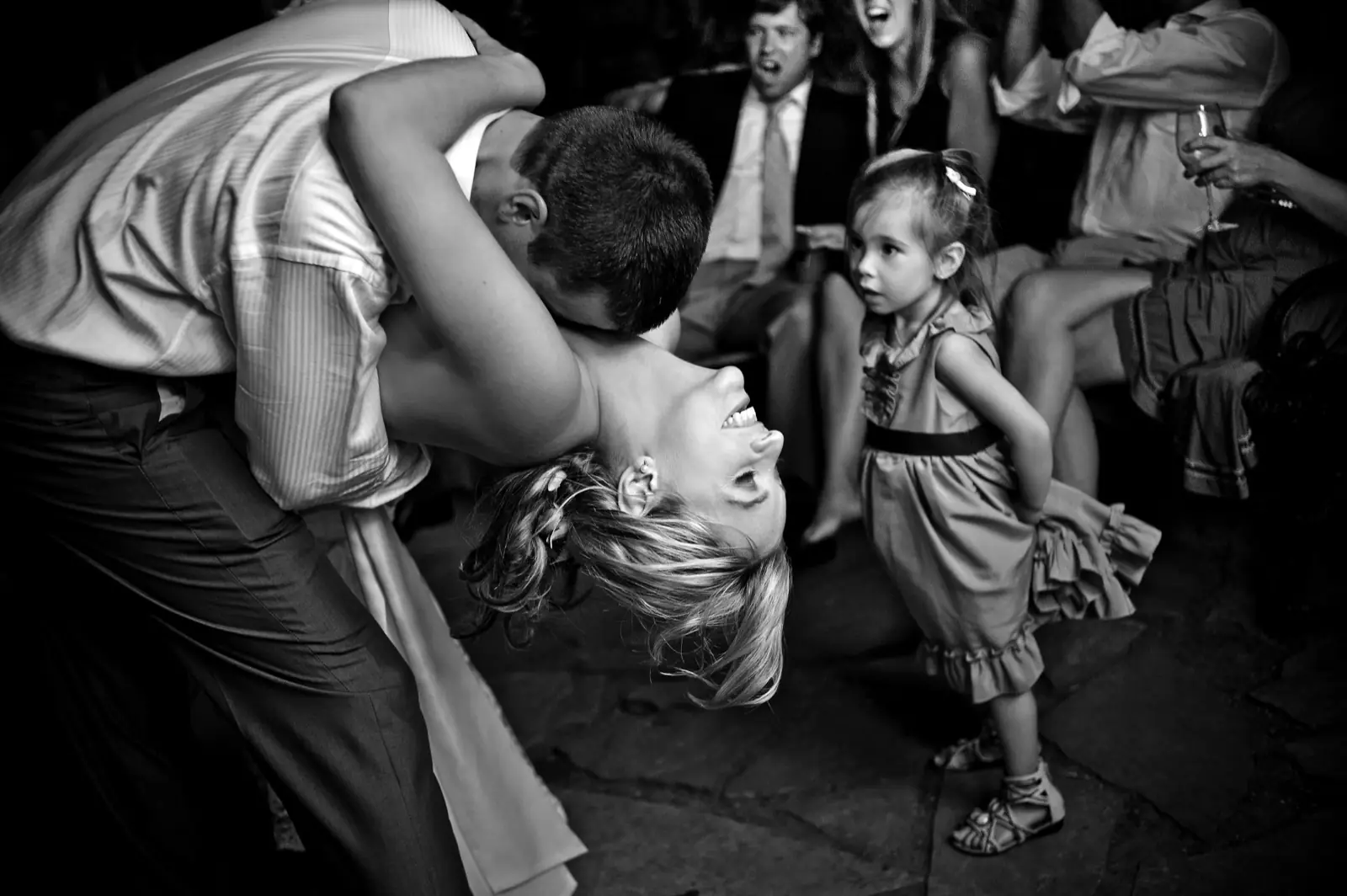 A bride dipped backwards laughing on the dance floor while a small girl watches, in black and white