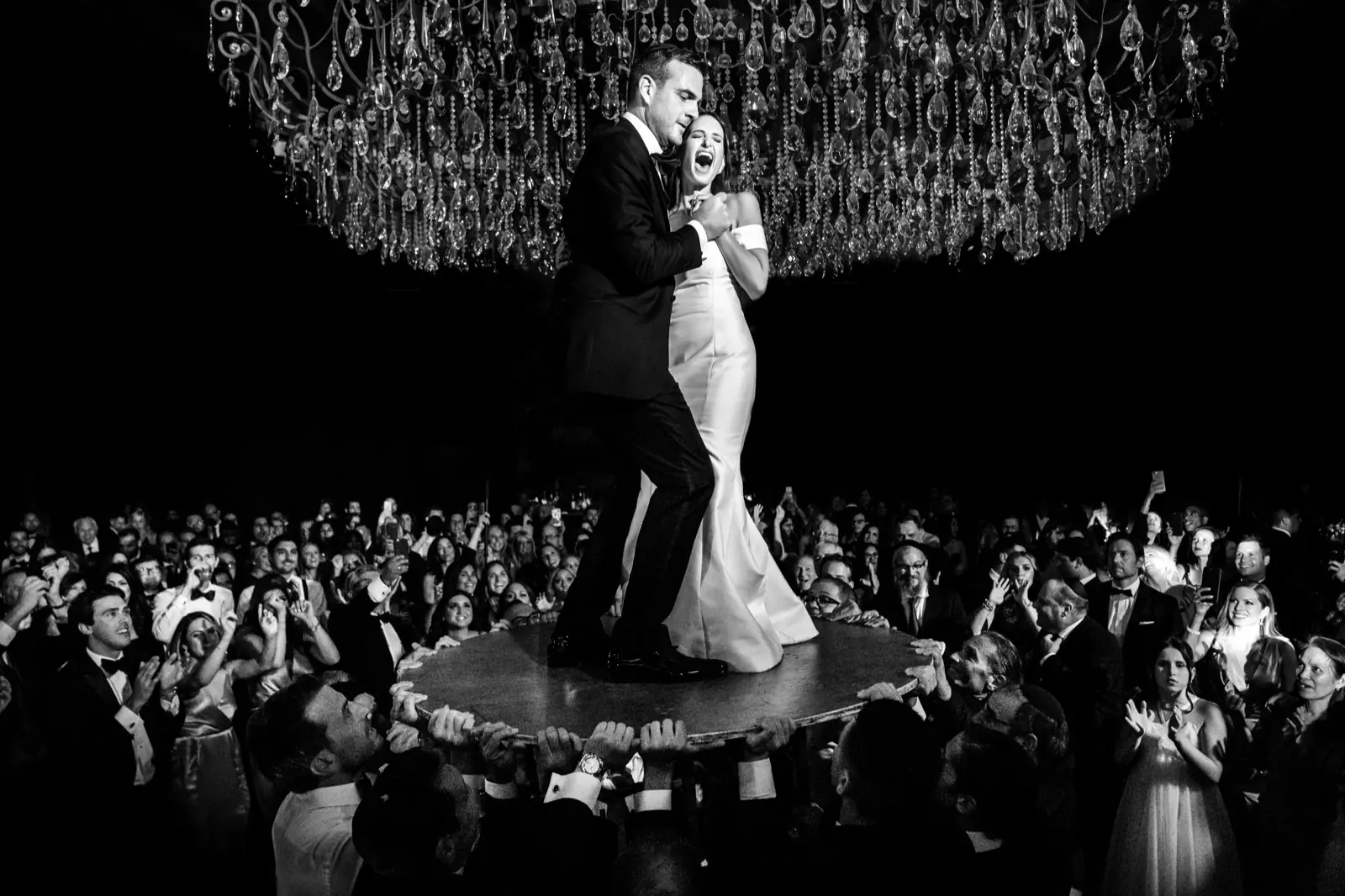 A bride alone on the dance floor under a chandelier, in black and white