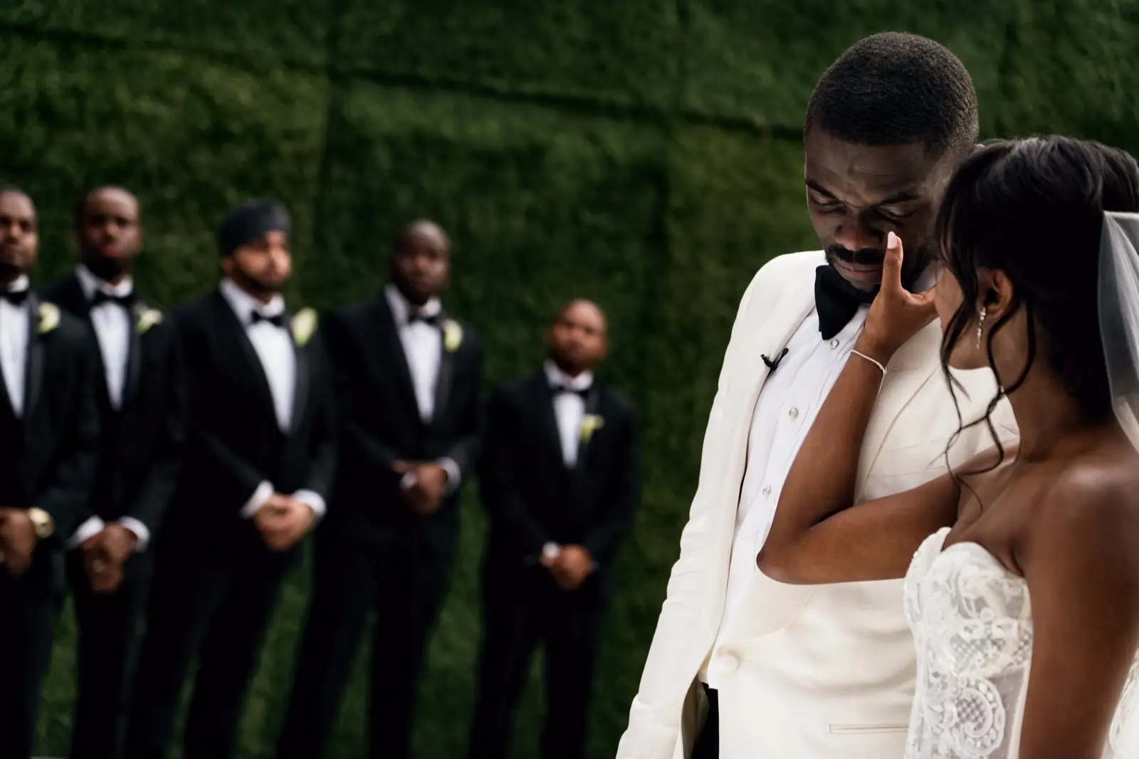 A groom with his hand to his face beside his bride, his groomsmen lined up on the grass behind