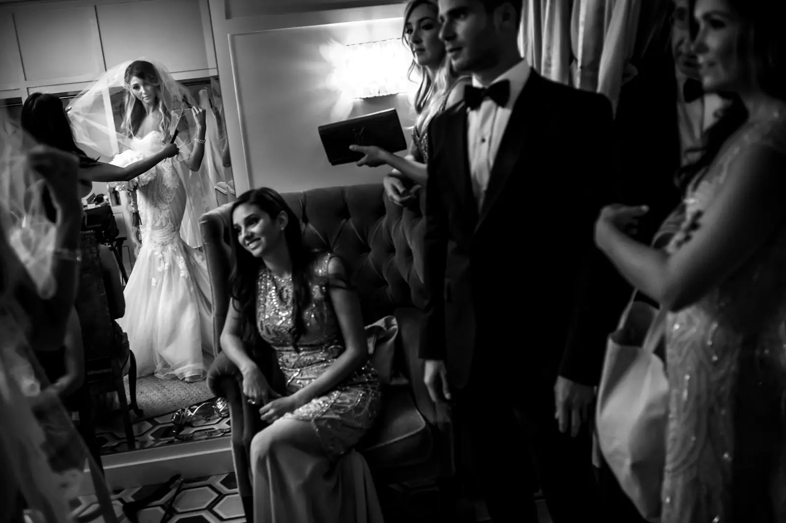 A crowded room before the ceremony, the bride in her dress at the edge of the frame, in black and white
