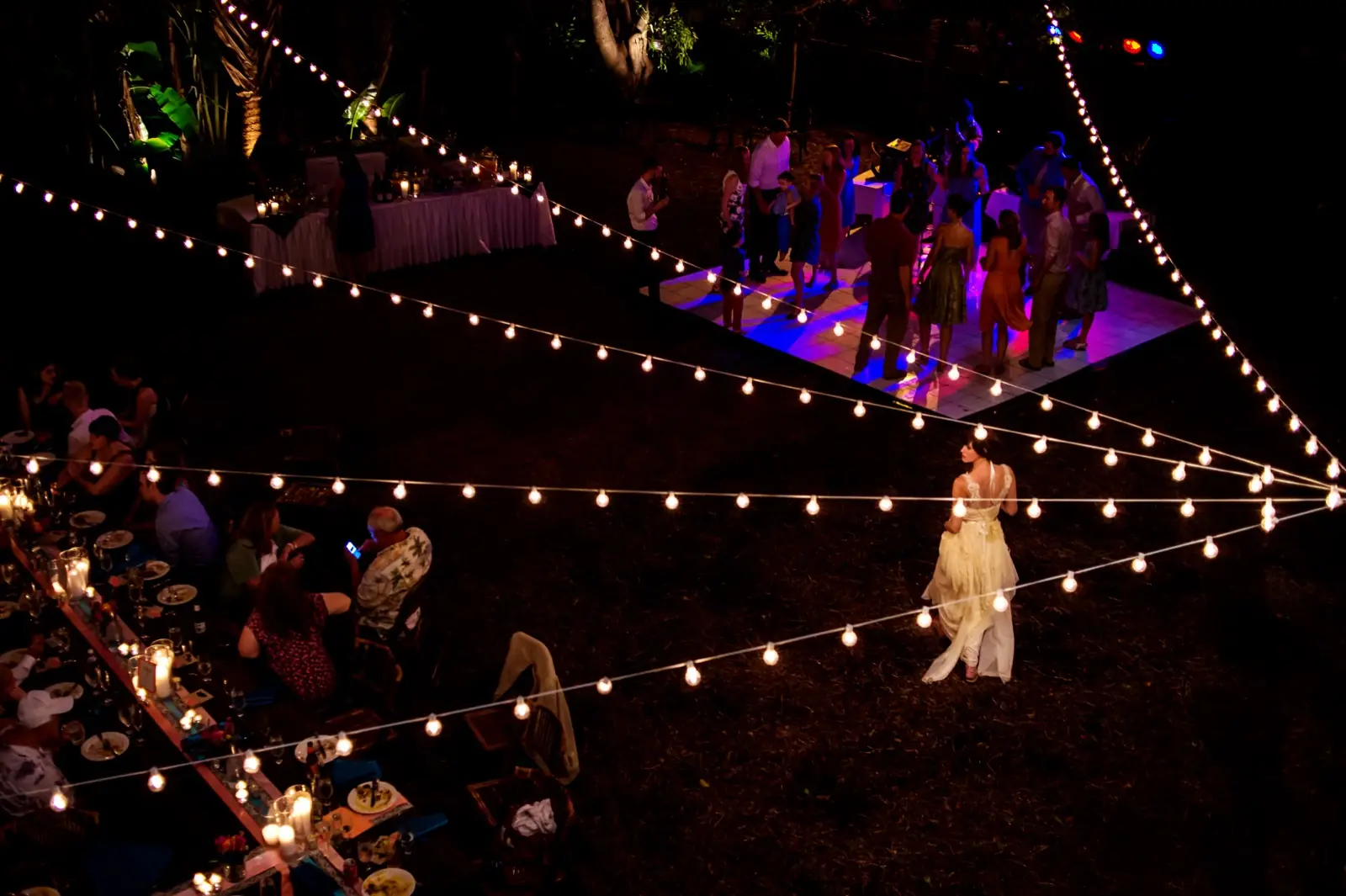 Strings of lights over the tables at night, a woman in a pale dress walking below