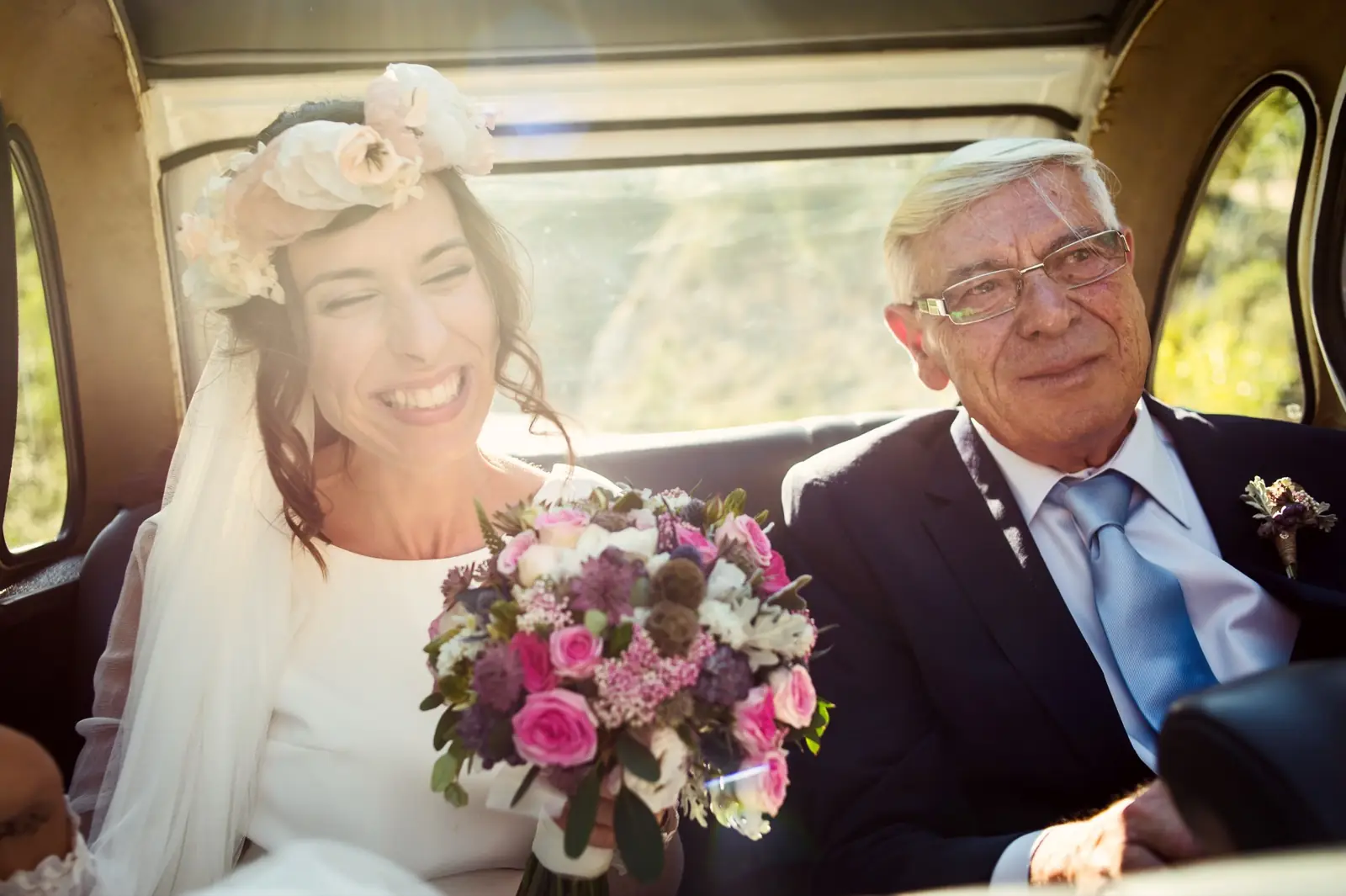 A bride in the back of a car beside her father, both smiling