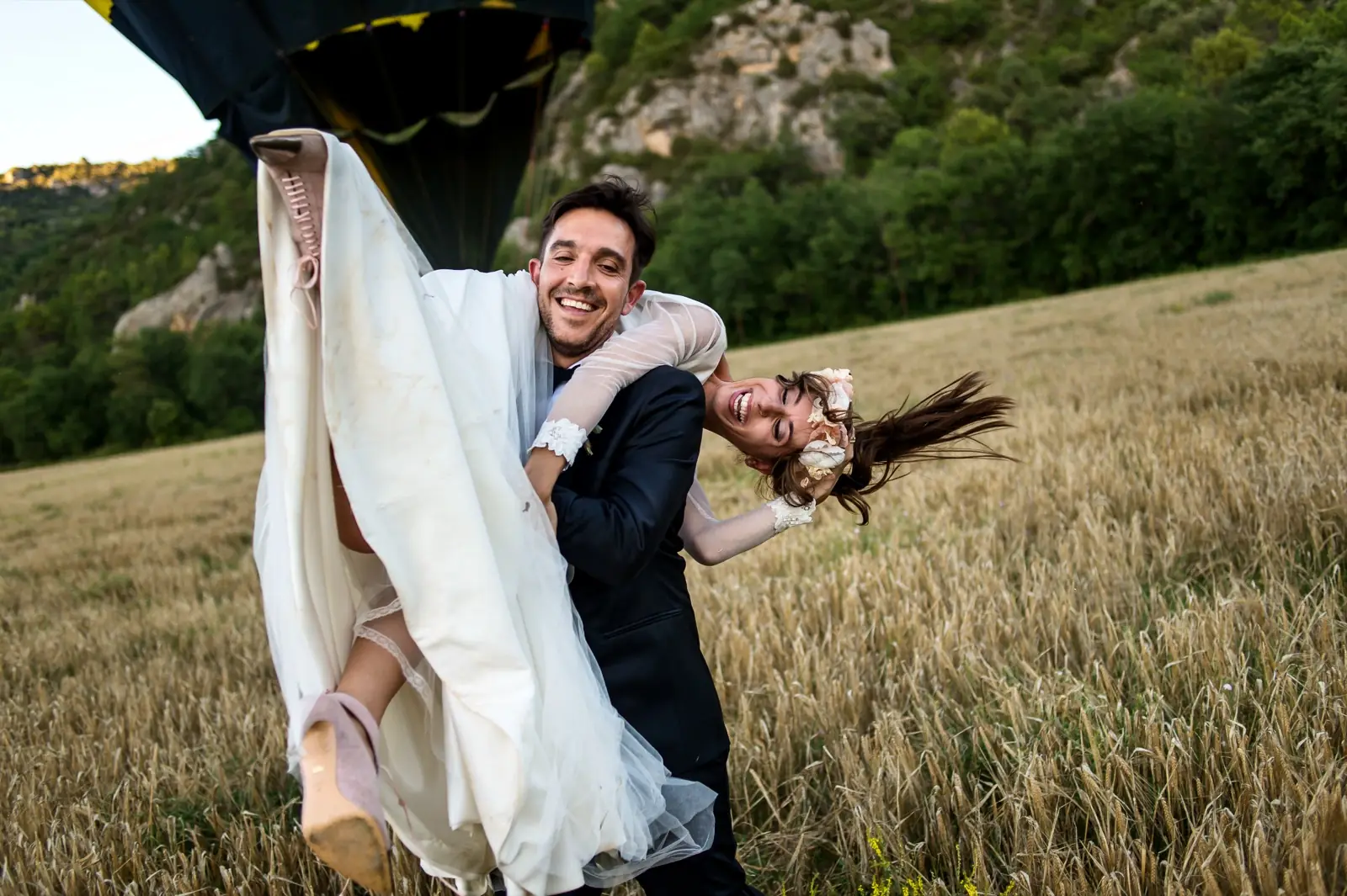 A groom carrying his laughing bride through a dry meadow, hills behind them