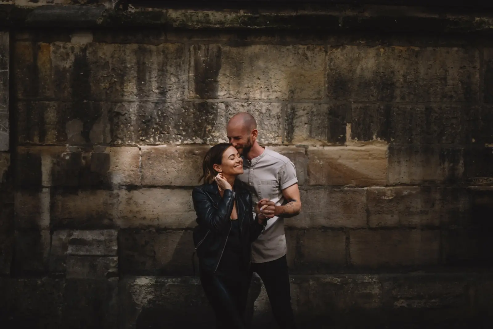 A couple standing against a weathered stone wall, her head at his shoulder