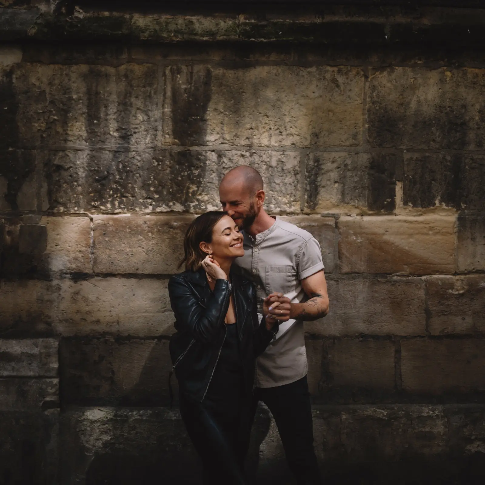 A couple close together against a streaked stone wall, both laughing