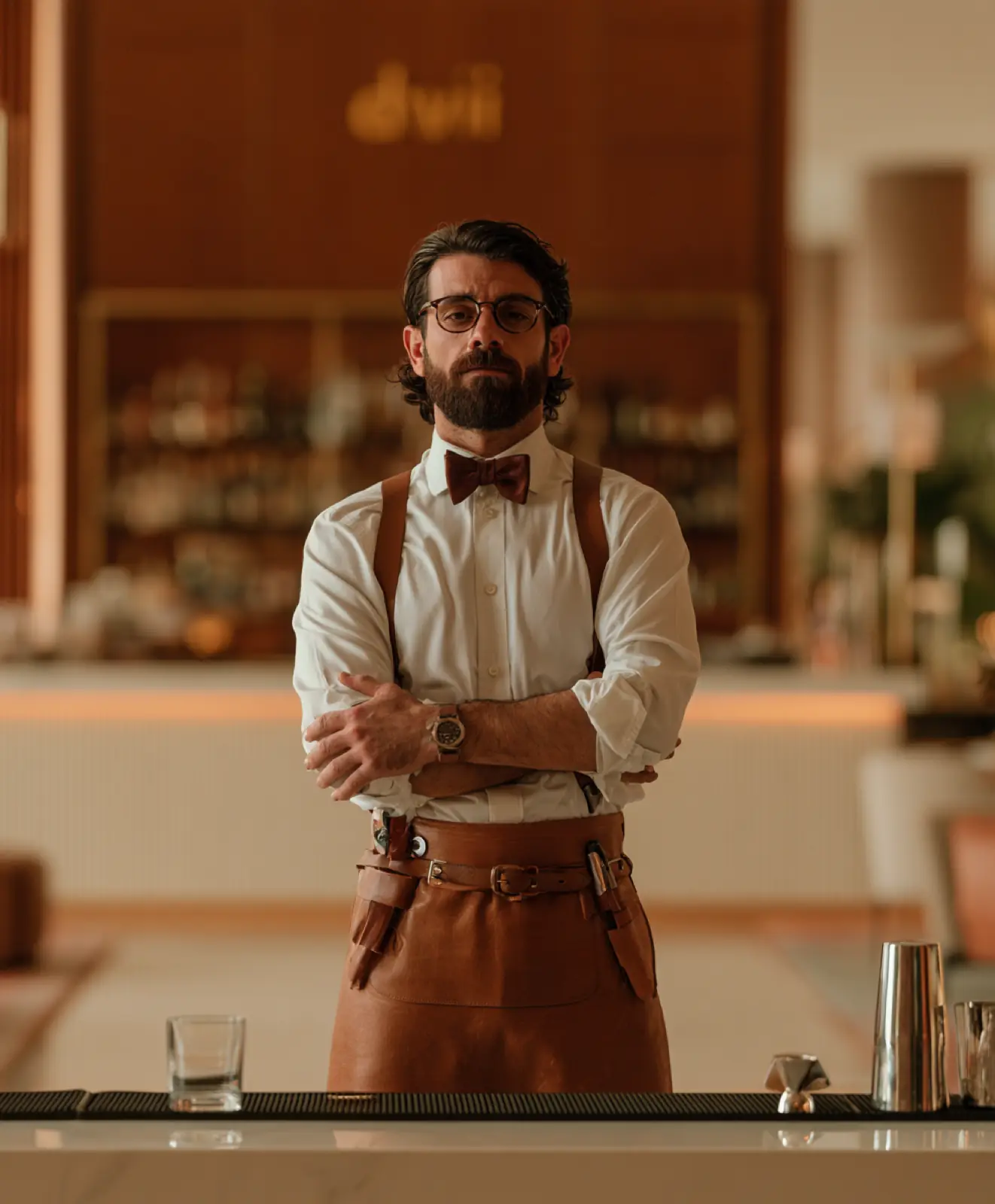 A barman in a leather apron and bow tie, arms crossed behind the bar